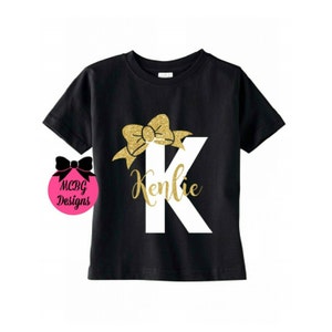 Op de afbeelding: Zwart T-shirt met een witte letter K en een gouden glitter strik erboven. De naam Kenlie staat onder de letter.