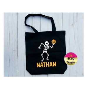 Puede incluir: Bolsa de lona negra con un esqueleto blanco sosteniendo una calabaza. La bolsa tiene el nombre "NATHAN" impreso en amarillo.