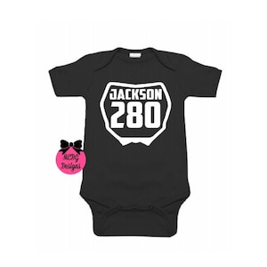 Puede incluir: Mono negro para bebé con un diseño de placa de número de carreras blanca. La placa dice "JACKSON 280".