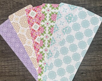May include: A collection of six decorative bookmarks with various floral and geometric patterns. The bookmarks feature a range of colors, including purple, gold, pink, green, and blue, and are arranged in a fan shape on a dark wooden surface.