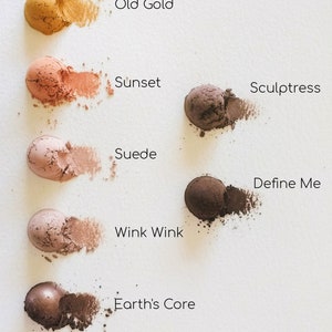 May include: Six eyeshadows in various shades of brown, pink, and gold. The eyeshadows are arranged in a vertical line from top to bottom. The names of the eyeshadows are written below each one: Old Gold, Sunset, Sculptress, Suede, Wink Wink, and Earth's Core.