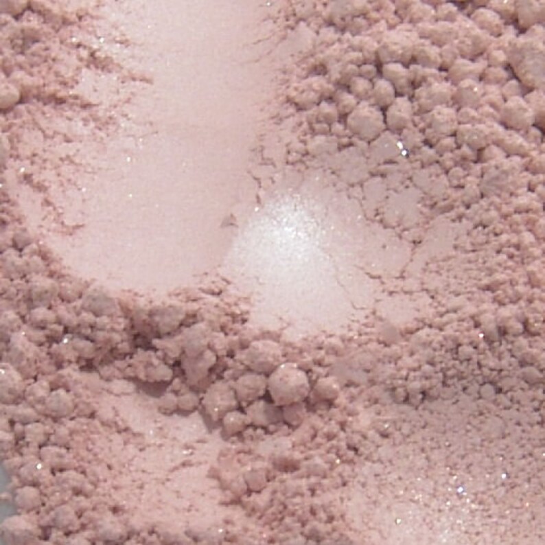May include: Close-up of a light pink, finely milled powder with a shimmering, pearlescent finish. The powder has a slightly textured appearance, with areas of concentrated pigment and lighter, more reflective patches.