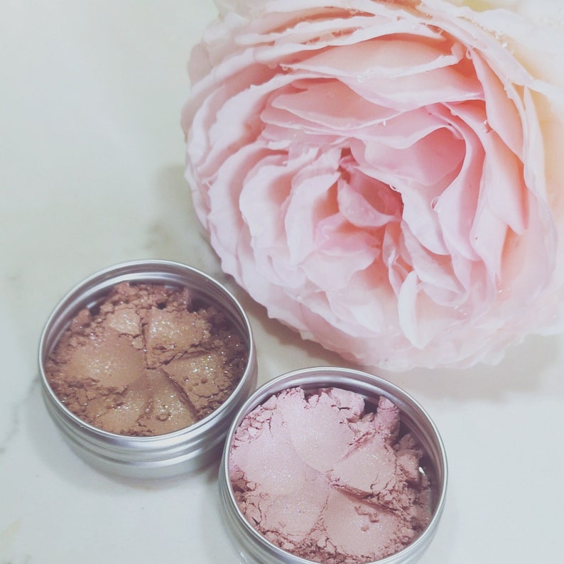 May include: Two small round containers with lids, filled with loose shimmery powder. One container holds a light brown powder, the other a light pink powder. A pink rose is in the background.