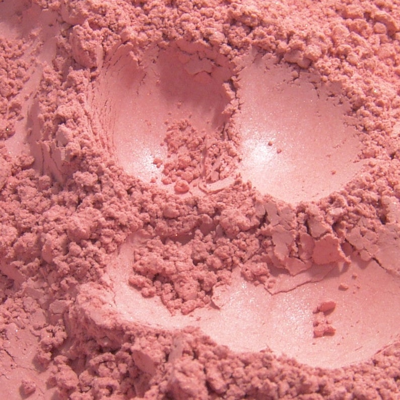May include: Close-up of a pile of pink powder with a slightly shimmery finish. The powder has a textured, granular appearance with smooth, compacted areas. The color is a soft, muted pink.