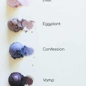May include: Four eyeshadow samples in shades of purple, blue, and black. The shades are labelled "Lilac", "Eggplant", "Confession", and "Vamp".