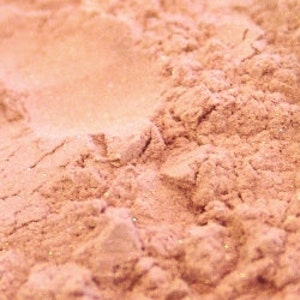 May include: Close-up of a pile of finely milled, light peach-colored powder with a subtle shimmer. The powder has a soft, textured appearance, with small particles visible. The overall effect is delicate and slightly iridescent, suggesting a cosmetic product.