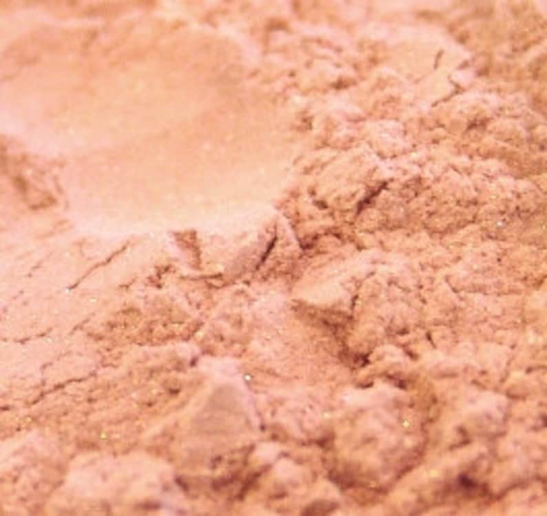 May include: Close-up of a pile of finely milled, light peach-colored powder with a subtle shimmer. The powder has a soft, textured appearance, with small particles visible. The overall effect is delicate and slightly iridescent, suggesting a cosmetic product.