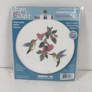 May include: A Learn a Craft counted cross stitch kit featuring a design of two hummingbirds and pink trumpet flowers on white Aida fabric inside a 6-inch plastic hoop. The packaging includes cotton thread, a needle, and instructions.