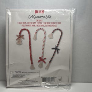 May include: A macrame kit in a clear plastic package. The kit includes materials to make three candy cane decorations. The candy canes are red, white, and black with decorative bows. The package text reads "Macrame Kit".