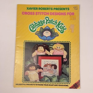 May include: A vintage book titled "Xavier Roberts Presents Cross Stitch Designs for Cabbage Patch Kids." The cover is yellow with green and red accents, featuring Cabbage Patch Kids dolls and a cross-stitch design. The book is a guide for cross-stitch projects.