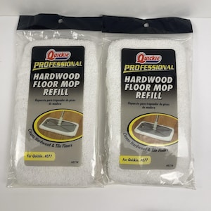 May include: Two white hardwood floor mop refills in clear plastic packaging. Each package features a black and gold label with the text "Quickie Professional Hardwood Floor Mop Refill". The label also includes an illustration of a mop and the text "Cleans Hardwood & Tile Floors".