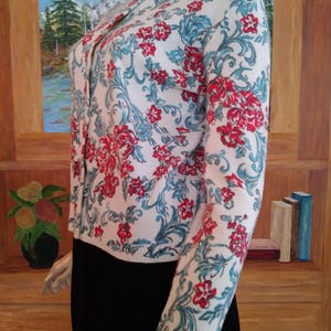 May include: A white cardigan with a floral pattern in red and teal. The long-sleeved sweater has a button-up front and a high neckline. The floral design covers the entire sweater, creating a vintage aesthetic.