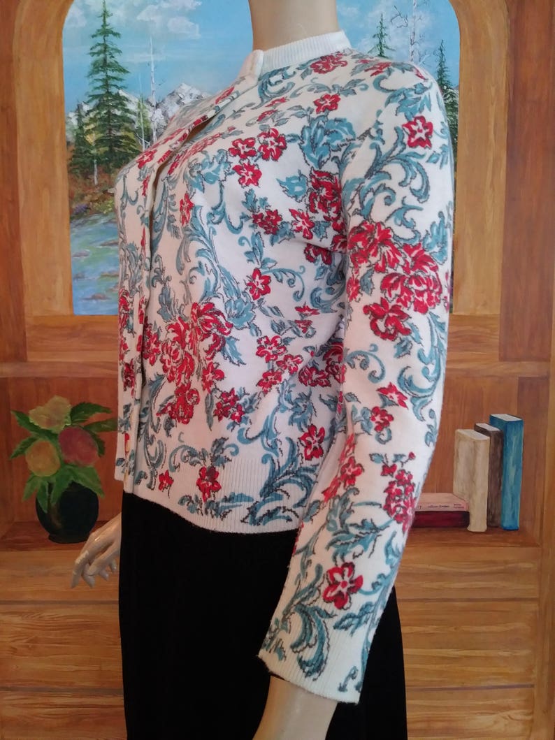 May include: A white cardigan with a floral pattern in red and teal. The long-sleeved sweater has a button-up front and a high neckline. The floral design covers the entire sweater, creating a vintage aesthetic.