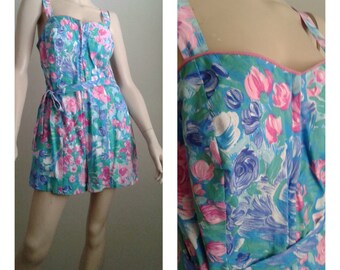 May include: A floral print romper with a pink trim, featuring a blue, green, and pink floral pattern. The romper has a tie at the waist and adjustable straps.
