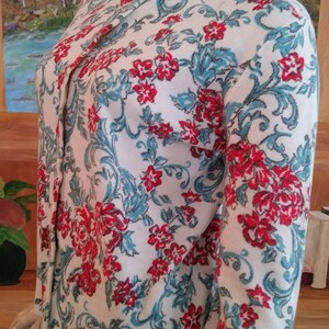 May include: Cream-colored cardigan sweater with a floral pattern in red and teal. The sweater has a round neckline and long sleeves. The floral design features stylized flowers and leaves. The sweater is buttoned up and appears to be made of a soft knit material.