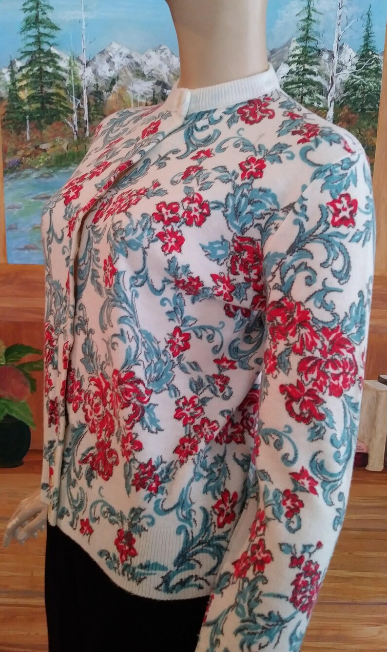 May include: Cream-colored cardigan sweater with a floral pattern in red and teal. The sweater has a round neckline and long sleeves. The floral design features stylized flowers and leaves. The sweater is buttoned up and appears to be made of a soft knit material.