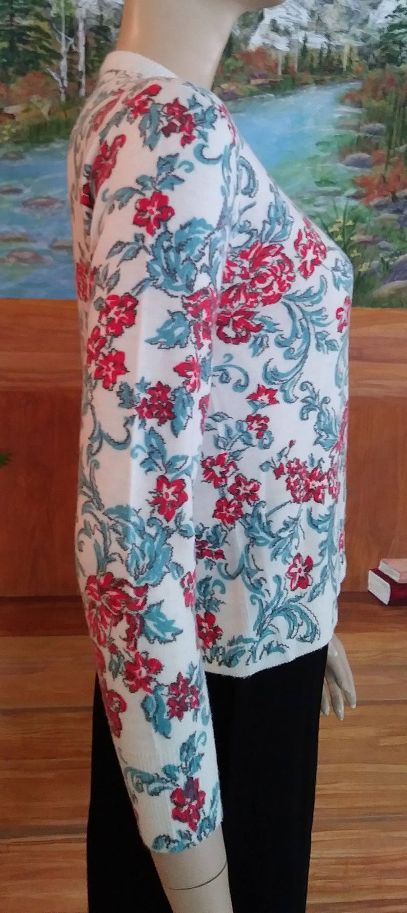 May include: White cardigan sweater with a floral pattern in red and teal. The sweater features a crew neck and long sleeves. The floral design includes stylized flowers and scrolling vines. The sweater is shown on a mannequin.