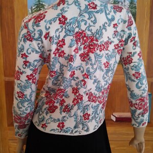 May include: White cardigan sweater with a floral pattern in red and teal. The sweater has long sleeves and a round neckline. The floral design covers the entire sweater, creating a vintage aesthetic. The sweater is paired with a black skirt.