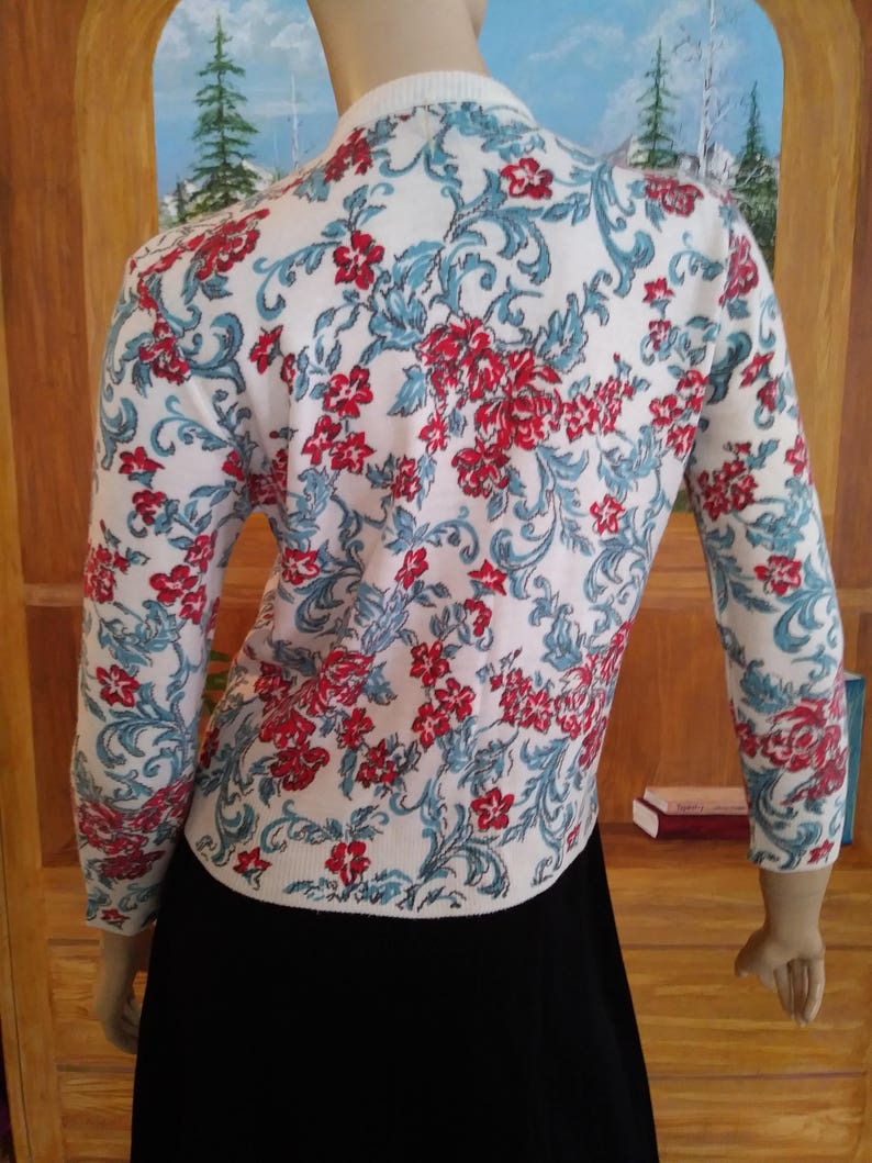 May include: White cardigan sweater with a floral pattern in red and teal. The sweater has long sleeves and a round neckline. The floral design covers the entire sweater, creating a vintage aesthetic. The sweater is paired with a black skirt.