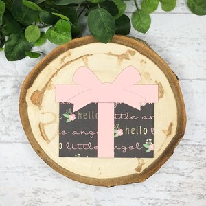 May include: A pink gift box with a bow on top. The box is decorated with a black and white pattern that says "hello little angel" in gold lettering.