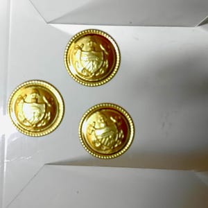 Puede incluir: Tres botones dorados con un diseño en relieve detallado. Los botones son redondos con un borde decorativo y un emblema central. Los botones están dispuestos sobre una superficie blanca.