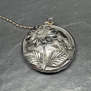 May include: A round silver-toned metal pendant featuring a raised relief design of a sunflower with two large leaves. The pendant is attached to a thin silver-toned ball chain. The surface of the metal has a textured, oxidised finish.