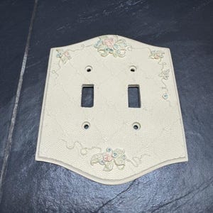 May include: A cream-colored double light switch wall plate featuring a textured honeycomb pattern and raised floral accents in soft pink and green. The rectangular plate includes two vertical rectangular cutouts and four screw holes.