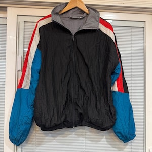May include: A black zip-up windbreaker jacket featuring a high collar with a grey interior lining. The sleeves are color-blocked with vibrant red, white, and blue panels. The jacket has a crinkled texture and a full-length front zipper closure.