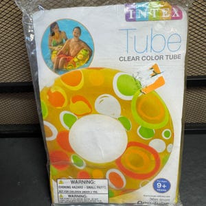 Op de afbeelding: Een transparante Intex zwemband in de originele plastic verpakking. De opblaasbare ring heeft een gele basis met een patroon van oranje, groene en witte cirkels. De verpakking bevat producttekst, een waarschuwingslabel en 91cm.