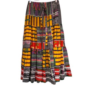 May include: A long, tiered skirt with a patchwork design. The skirt features a mix of patterns, including stripes, geometric shapes, and floral motifs, in a color palette of yellow, orange, purple, and black. It has a drawstring waist and a flowing silhouette.