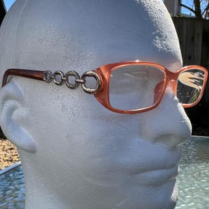May include: Peach-colored eyeglasses with a rectangular frame and clear lenses. The temples are a darker brown and connect to the frame with a series of silver-toned, circular embellishments. The glasses are displayed on a white mannequin head.