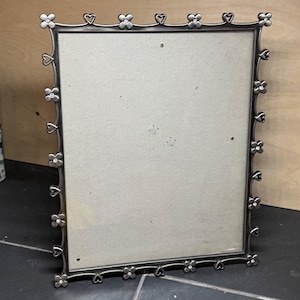 Può includere: Una cornice rettangolare color argento con un design decorativo floreale e a forma di cuore. Il bordo della cornice presenta forme alternate di fiori e cuori, con piccoli abbellimenti sui fiori. La cornice è vuota, con un retro di colore neutro.