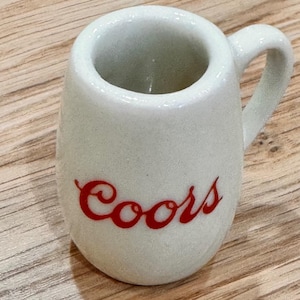 May include: A small, off-white ceramic mug with a handle. The word "Coors" is written in red script on the front of the mug. The mug has a rounded body and a slightly flared rim.
