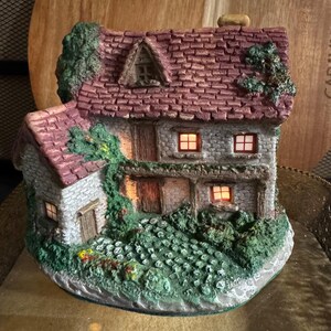 May include: A miniature cottage figurine with a textured red shingled roof, stone-patterned walls, and green foliage accents. The structure features multiple windows with warm interior lighting, wooden-style doors, and a cobblestone-textured base.