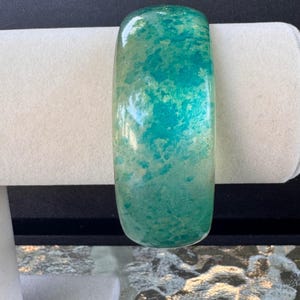 Könnte beinhalten: Ein breites, oval geformtes Armreif-Armband. Das Armband ist eine Mischung aus Türkis und hellgrünen Farben mit Marmoreffekt. Das Armband wird auf einem weißen, zylindrischen Ständer präsentiert.
