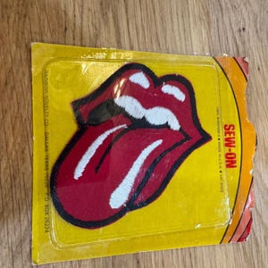 May include: A red and white embroidered patch in the shape of a mouth with a tongue, packaged in a yellow plastic blister pack. The package has the words "SEW-ON" printed on it. The patch is made in the USA.