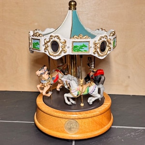 May include: A decorative carousel music box with a wooden base and a white and teal canopy. The carousel features several colorful horses with gold accents. The base has a gold emblem.