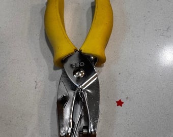 May include: A silver-colored hole punch with yellow handles. The handles are curved and padded for grip. The hole punch is made of metal and has a shiny finish. A small red star is next to the hole punch.