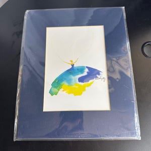 May include: Watercolor painting of a figure in a blue, yellow, and green dress with a white heart above. The artwork is in a white mat and a dark blue frame. The artist's signature, "leahjoy", is in the lower right corner.