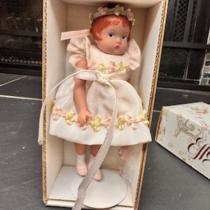 Puede incluir: Una muñeca vintage con cabello rojo, vestida con un vestido rosa claro con detalles florales, se exhibe en su caja de cartón original. La muñeca tiene una delicada corona floral y está colocada sobre un soporte blanco. Se ve una caja con ilustraciones.