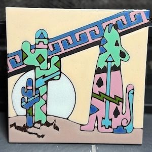 May include: A square ceramic tile featuring a desert scene with a stylised coyote and cacti in shades of mint green, pink, and blue against a cream background. A diagonal blue and pink geometric border crosses the upper portion of the tile.