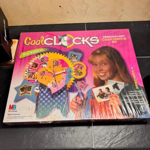 May include: A pink boxed Cool CLOCKS personalized clock-making kit. The box features a colorful clock design with photo frames and the text "You Build It". Includes 7 fun-shaped frames to hold photos. Ages 8 and up.