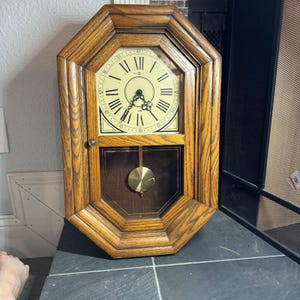May include: A wooden wall clock with an octagonal frame. The clock face is cream-colored with black Roman numerals and hands. A pendulum swings below the clock face. The clock is made of wood with a natural finish.