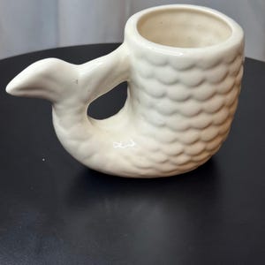 May include: Cream-colored ceramic mug shaped like a mermaid. The mug features a tail as the handle and a textured, scale-like pattern on the body. The tail curves to form the handle, with an opening in the middle.