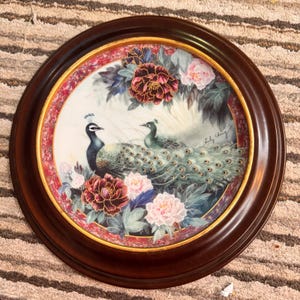 Op de afbeelding: Ronde decoratieve plaat met een donkerbruine houten lijst. De plaat toont een schilderij van twee pauwen, omringd door bloemen in roze, bordeauxrood en blauw. De plaat heeft een gouden en rode rand.