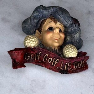 May include: A whimsical golf-themed figurine featuring a smiling character wearing a blue cap and holding two golf balls. A red banner below reads "Golf Golf Lie Golf". The figurine has a detailed, hand-painted appearance.