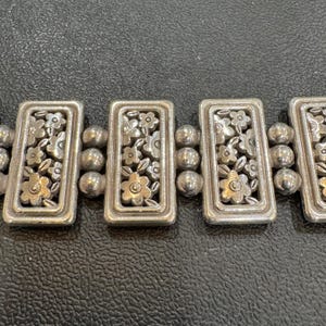 May include: Silver-toned bracelet with rectangular floral design links. Each link features a detailed flower pattern with a silver frame. The bracelet is composed of multiple rectangular links connected by small, round beads.