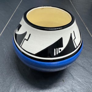 May include: A ceramic bowl with a rounded shape, featuring a blue base, white center with black geometric designs, and a black rim. The interior is a light yellow color. This decorative bowl is suitable for home decor.