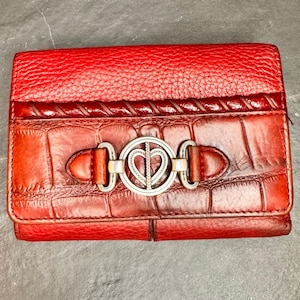 May include: Red leather wallet with a textured top and a crocodile-patterned bottom. The wallet features a silver-toned heart-shaped clasp and brown leather accents. The wallet is rectangular and has a bi-fold design.