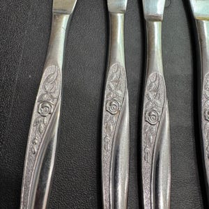 May include: Close-up of several silver knives. The handles feature a floral rose design. The knives are arranged in a row, with the blades facing upwards. The background is a dark, textured surface.